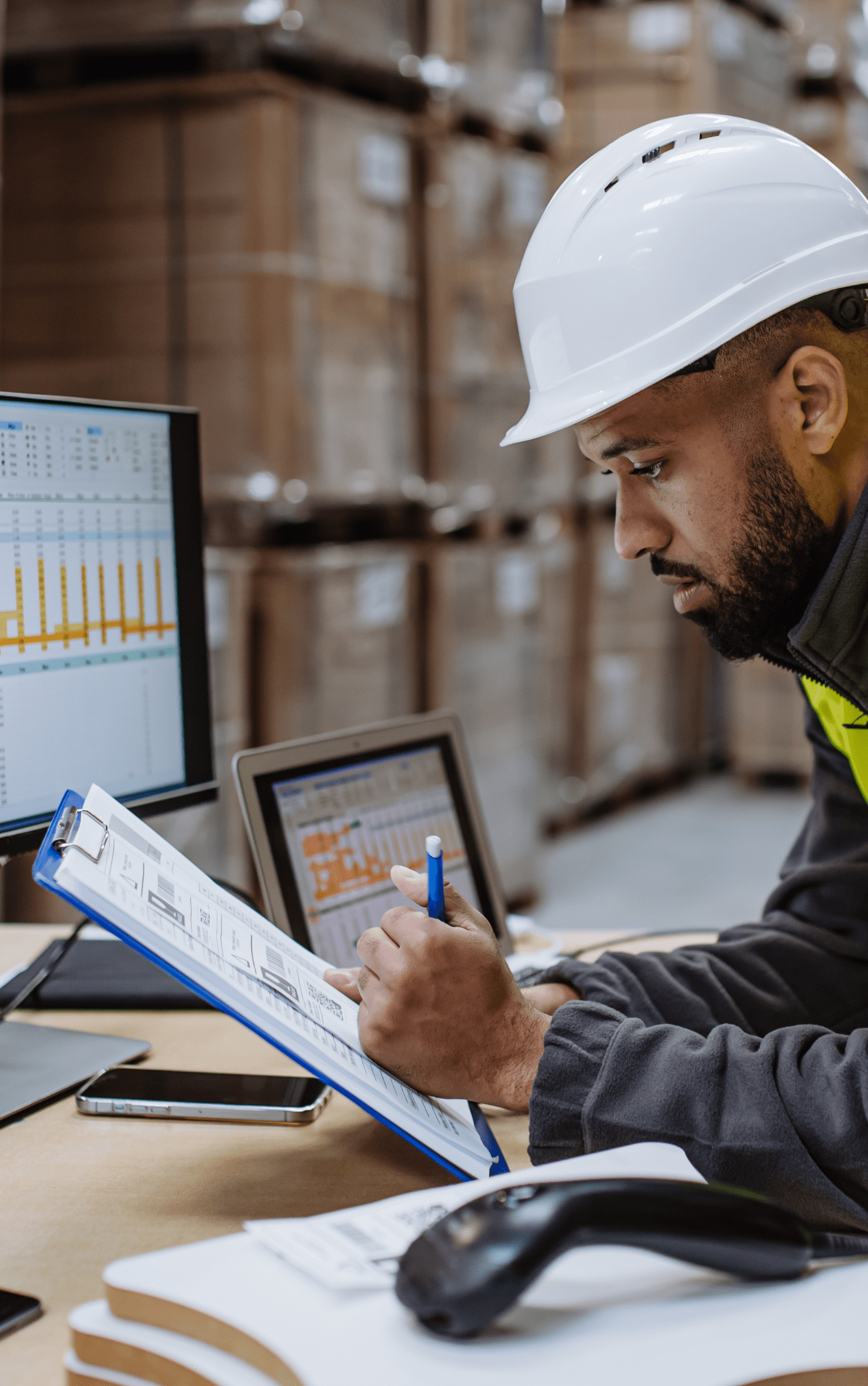 Man in a white hard hat looking over a clipboard and computer spreadsheet.