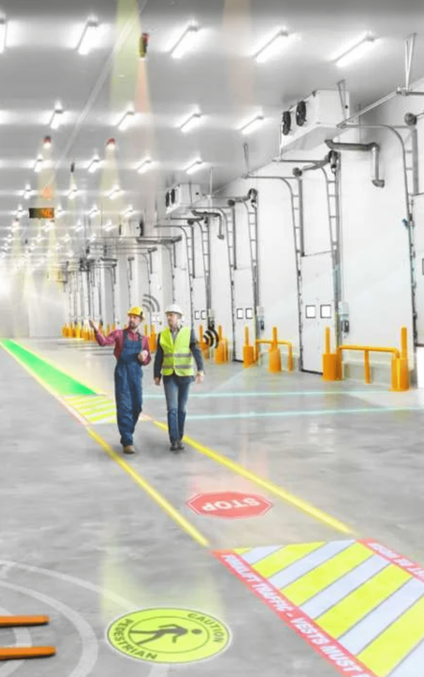 Two workers in reflective vests and hard hats walking in a warehouse environment.